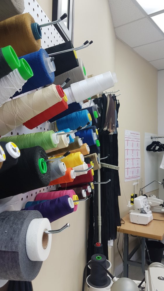 A pegboard wall of colourful thread spools beside a rack of garments and a long-arm sewing machine at New Stitch Tailor